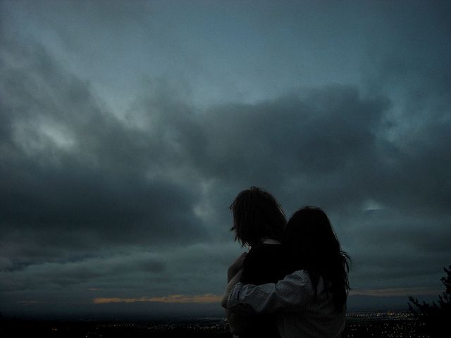 two people embracing each other in front of a dark sky with clouds and city lights behind them