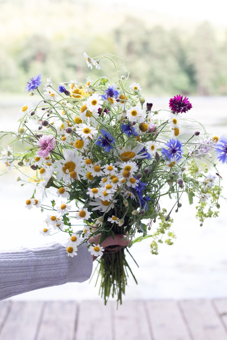 a person holding a bouquet of wildflowers and daisies on a wooden deck