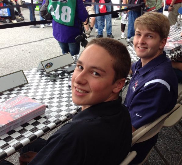 two young men sitting at a table with laptops on it and people in the background