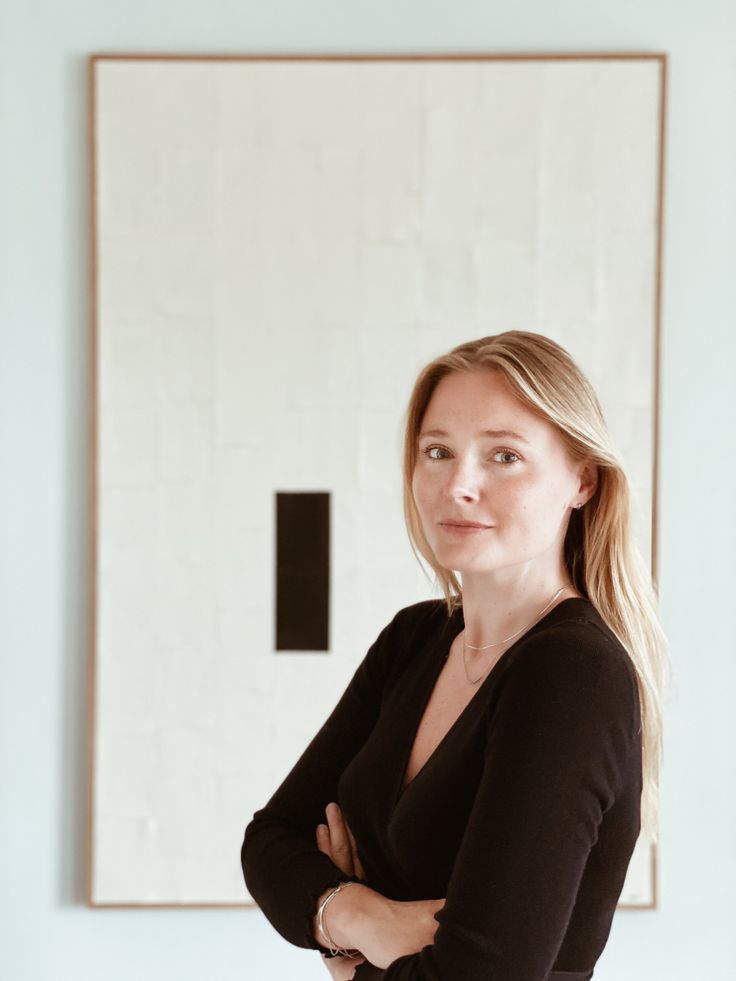 a woman standing in front of a white wall with her arms crossed and looking at the camera