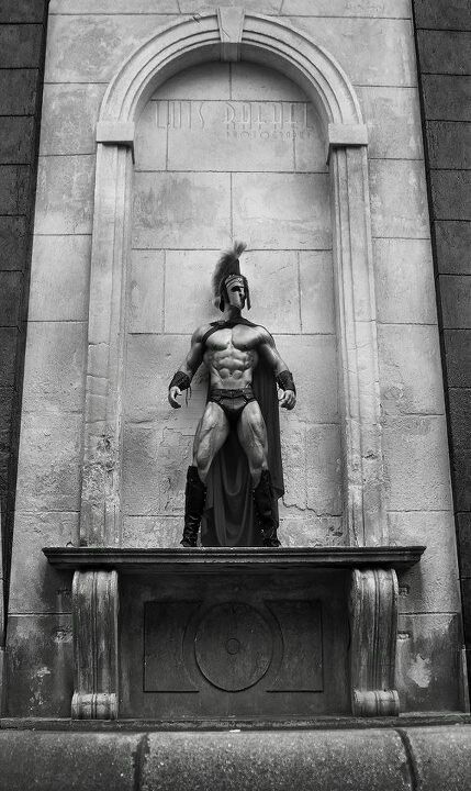 a statue of a man dressed as a roman warrior stands on a ledge in front of a stone building