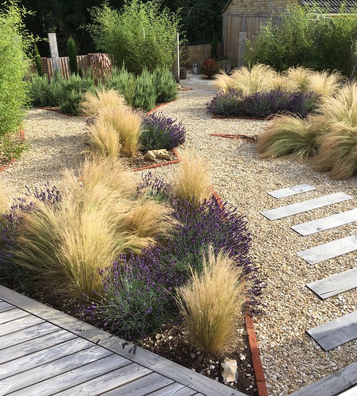 an outdoor garden with gravel, grass and flowers in the foreground is a wooden walkway that leads to a small building