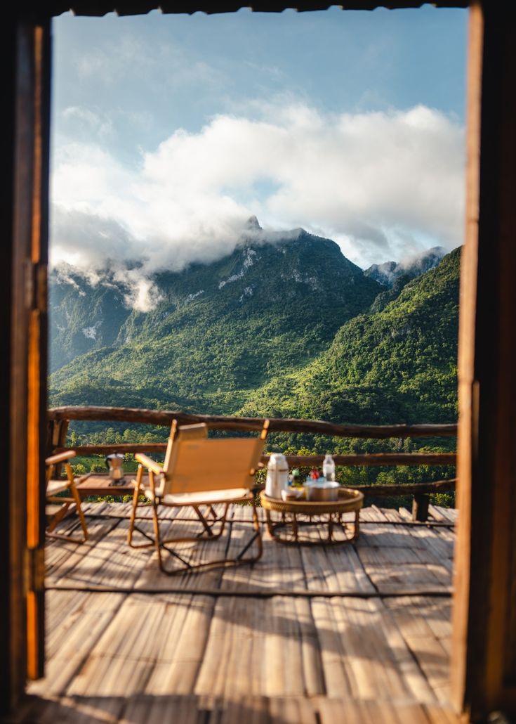 two chairs sitting on top of a wooden deck next to a table with drinks in front of mountains