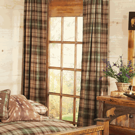 there is a bed with plaid sheets and pillows next to a wooden table in front of a window