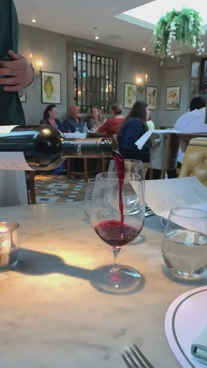 This may contain: a person pouring red wine into a glass at a table with people in the background