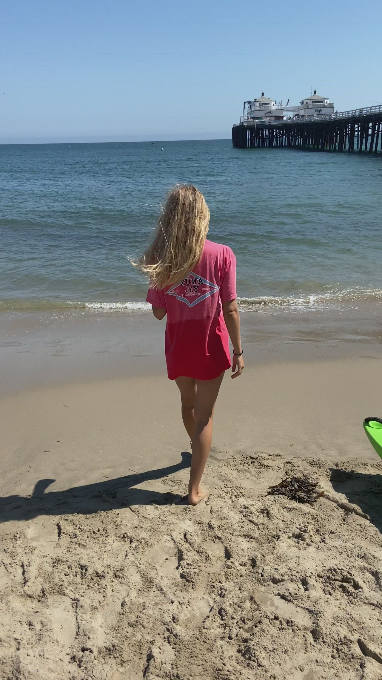 This may contain: a woman in a pink bathing suit and sunglasses standing on the beach with a pier in the background