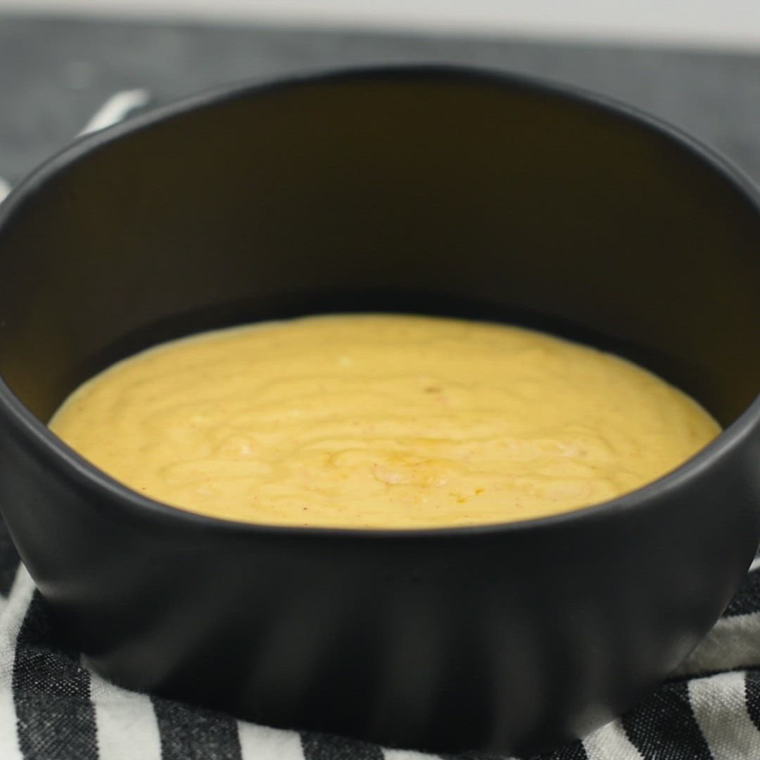 This may contain: a hand holding a tortilla chip over a black bowl filled with yellow dip