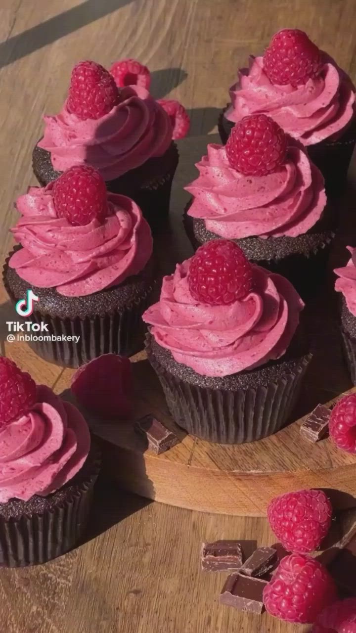 This may contain: chocolate cupcakes with pink frosting and raspberries arranged on a wooden platter