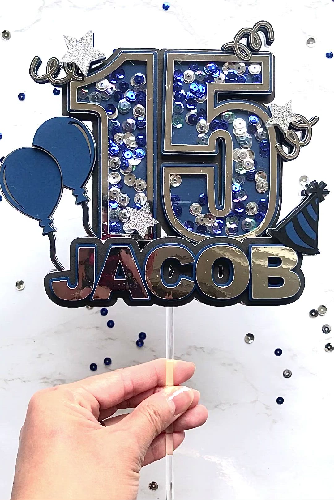 This may contain: a hand holding a blue and silver birthday cake topper