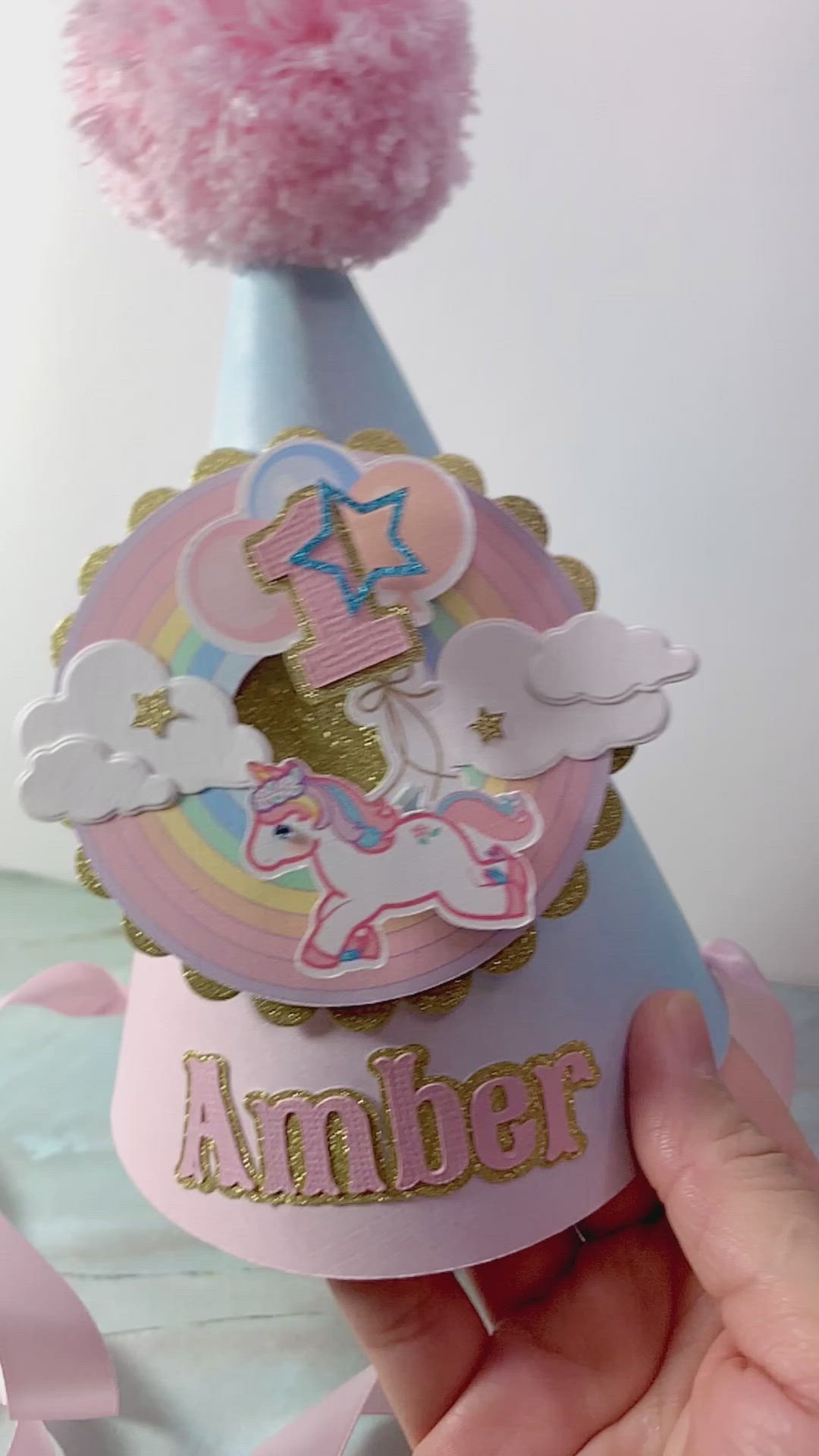 This may contain: a hand holding up a pink and blue party hat with an unicorn on the top