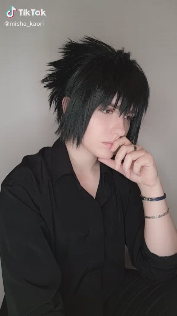 This may contain: a young man with black hair sitting in front of a gray wall and looking at the camera