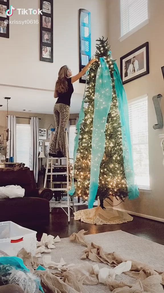 This may contain: a woman is standing on a ladder near a christmas tree in the middle of a living room