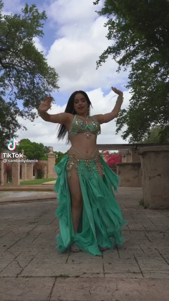 This may contain: a woman in a green belly dance outfit posing for the camera with her hands up