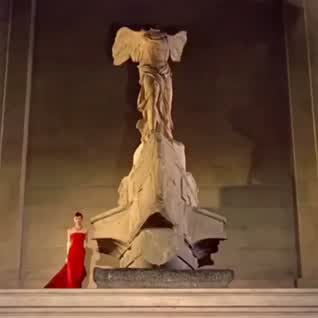 This may contain: a woman in a red dress standing on top of a white pillar with an angel statue behind her