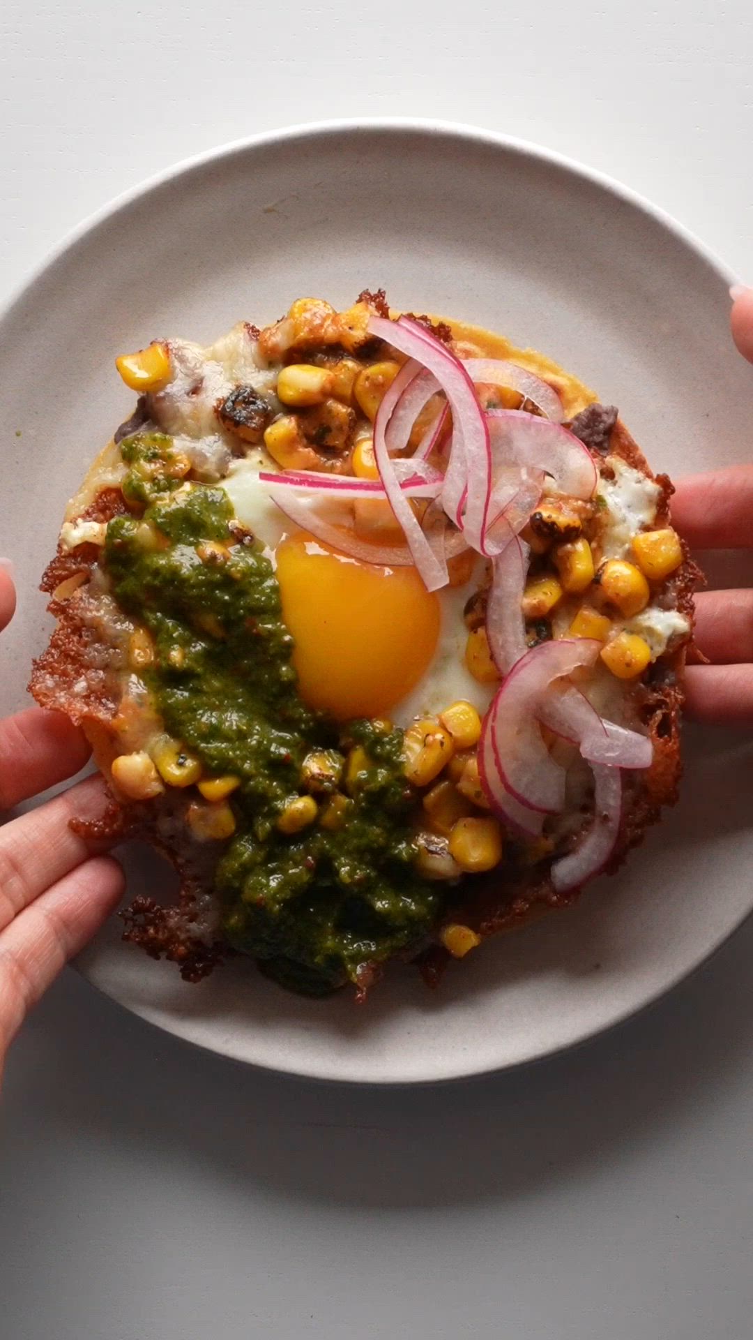 This contains: Making a crispy cheese ring in a pan and adding and egg in the middle with roasted corn, then spreading refried black beans on a tortilla and adding the egg with pickled onions and zhoug sauce.