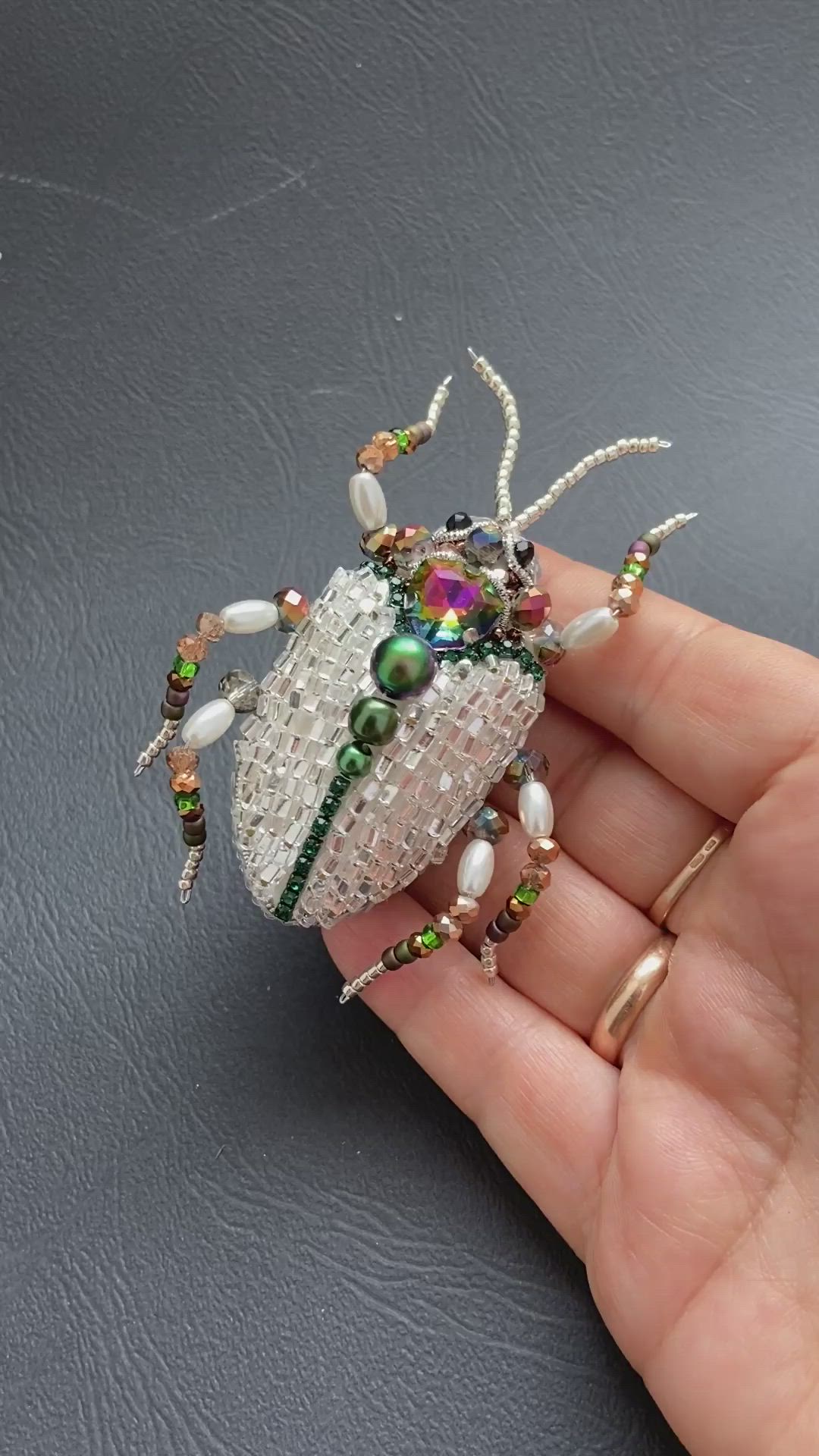 This may contain: a hand holding a small white and green bug brooch