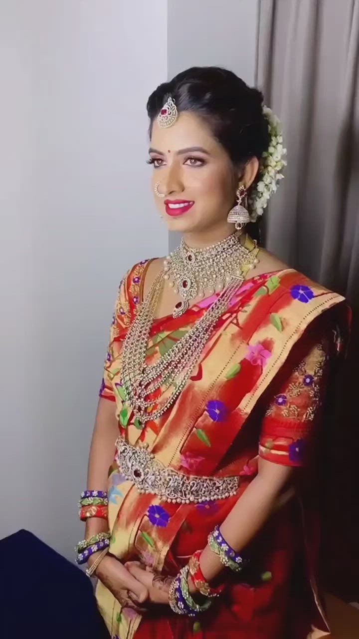 This may contain: a woman in a red and gold sari with jewelry on her neck, standing next to a window