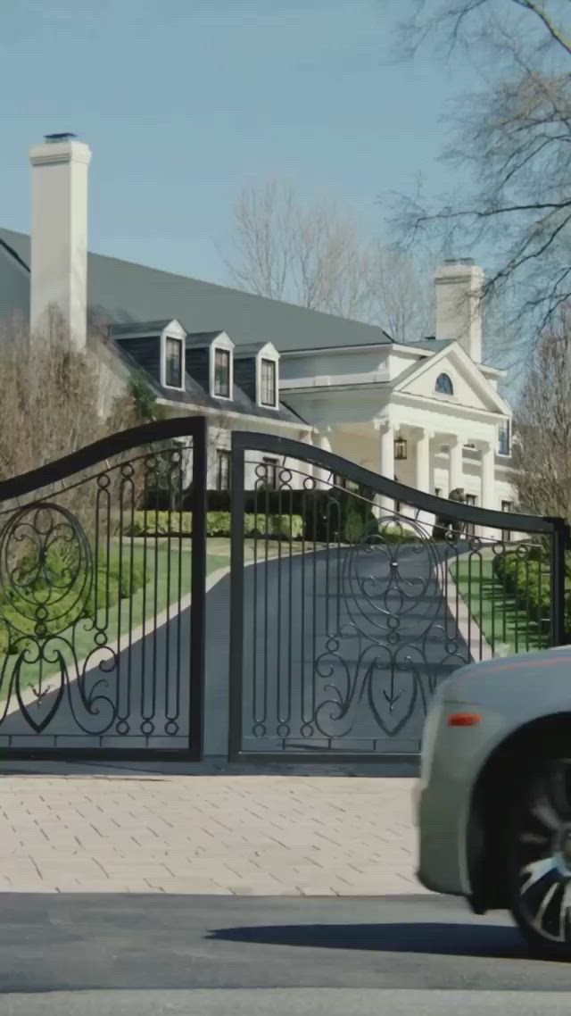 This may contain: a white car driving past a large house with a driveway and gate in front of it