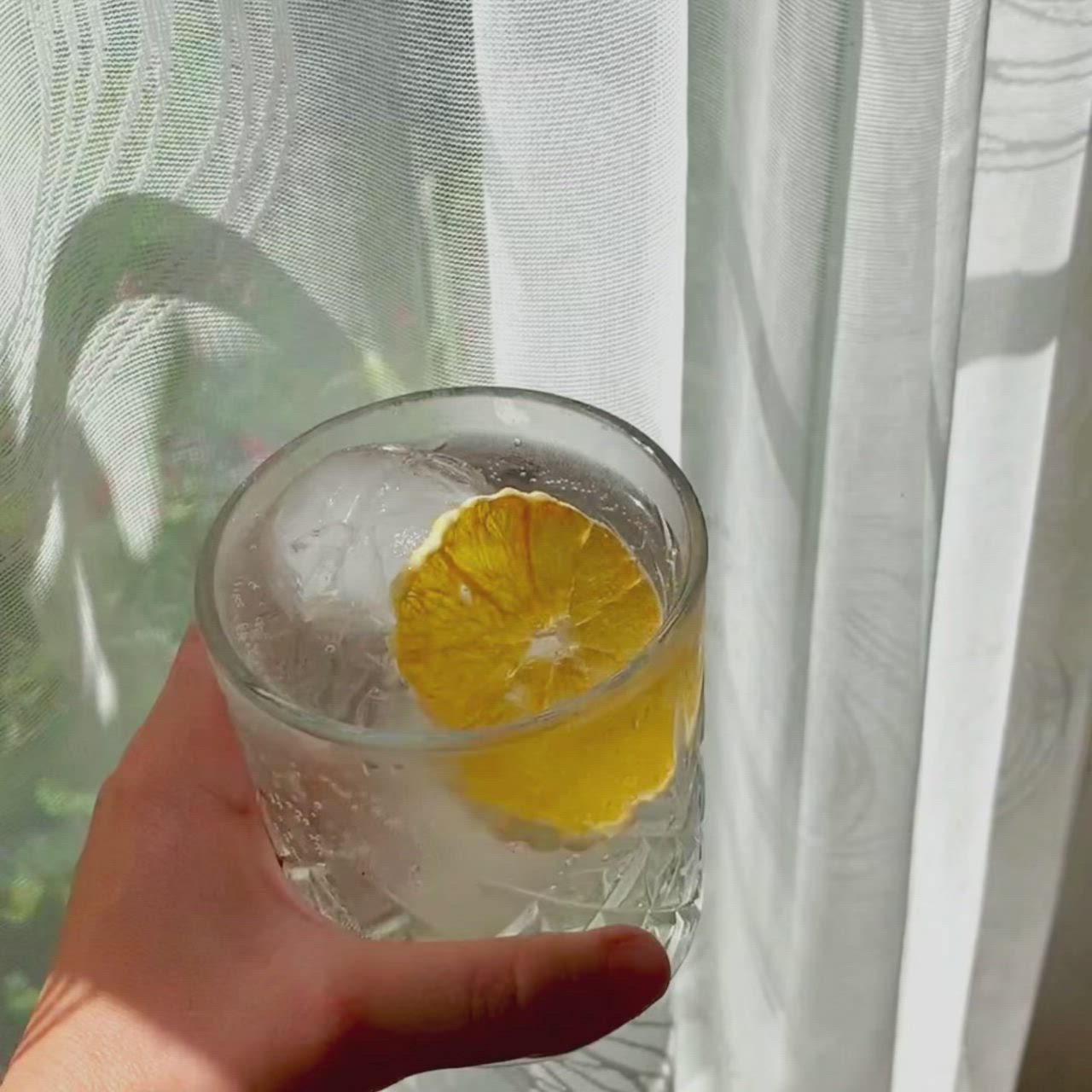 This may contain: a hand holding a glass with water and an orange slice in it, near a window