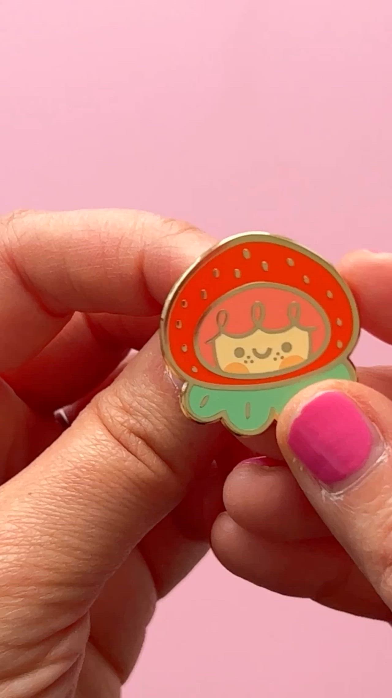 This contains: A hand showing a strawberry enamel pin. The pin has a girl face in the middle ad if she was wearing a strawberry hat with green leaves. The pin has a pink rubber clutch on the back to secure it to bags and jackets.