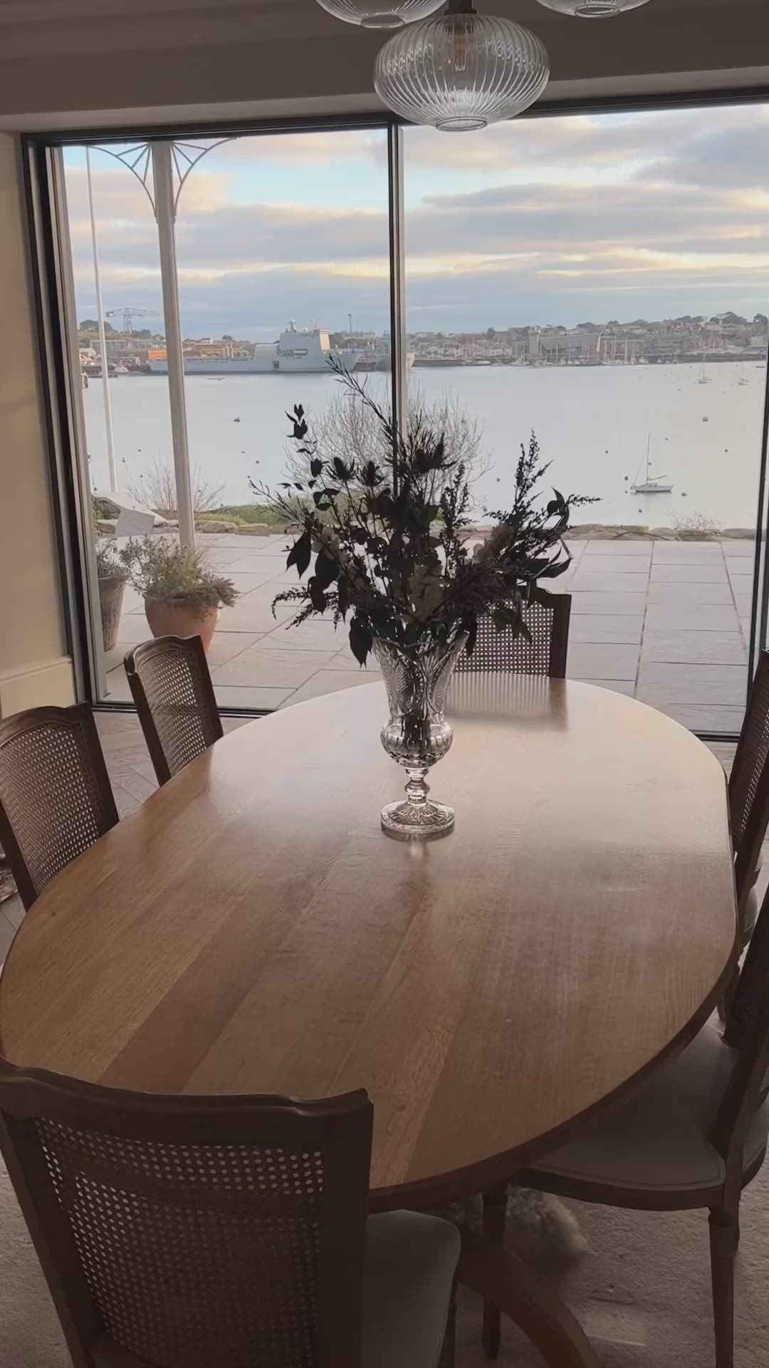 This may contain: a wooden table with chairs around it in front of a large window overlooking the water