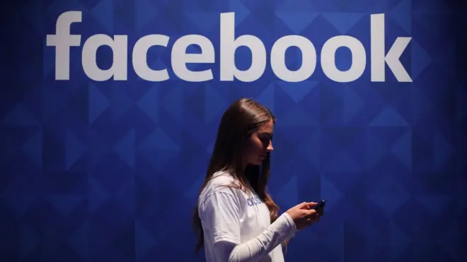 A woman uses her phone under a logo of the social media giant Facebook