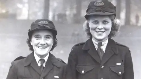 Ann Armitage and Betty Leverton looking at the camera. Anne has dark, short hair and wears a cap. Betty is taller and has curled blonde hair and wears a cap.