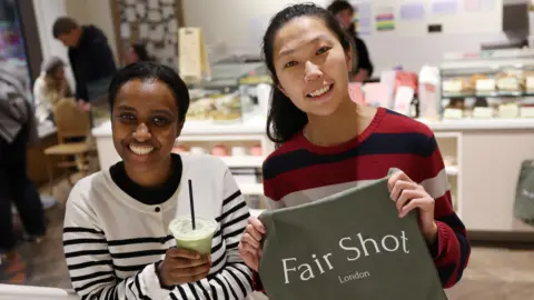 Two women holding a green tea towel with the words Fair Shot London and a smoothie. Both women are smiling at the camera.