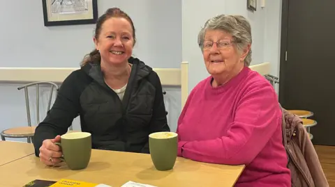 Kirsty Lawford and her Mum Margaret Blackshaw sit at a wooden table with two green cups in front of them. Kirsty is in her 50s and has red hair tied back and wears a black coat. Her mum Margaret is in her 80s and wears a bright pink jumper. 