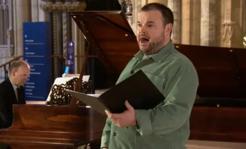 A man with short dark hair and dark-coloured stubble singing in a Church. He is wearing a long sleeve green shirt and is holding a folder open with his left hand. A man in a black suit is playing a piano behind him.