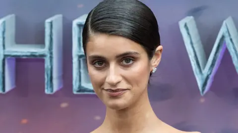 A woman with slicked back dark hair, parted to the side, stands in front of a hoarding on a red carpet. She's wearing expensive-looking diamond stud earrings.