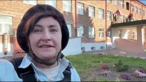 Journalist wearing a protective helmet speaks into the camera as troops behind her remove a Russian flag from a building.