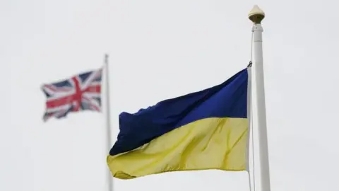 A Ukrainian flag flying on a pole against a grey sky. In the background there is a Union Jack flag flying out of focus.