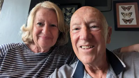 The image shows an elderly couple smiling, a woman on the left with short blonde hair and a bald man on the right. Behind them is a wall painted light blue with two picture frames containing pictured of three butterflied in each. The frame on the right contains orange and yellow butterflies, while the frame in the centre contains blue butterflies.