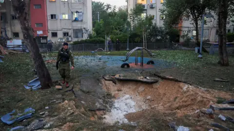 A man in a khaki uniform stands in a playground in front of a block of flats, looking down at a crater in the ground