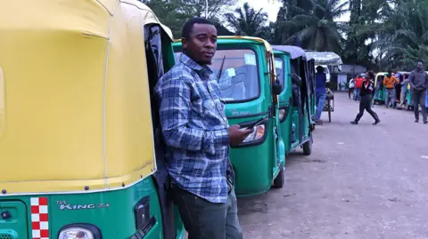 A man in jeans, sliders and a checked shirt stands by his three-wheeled vehicle as he waits to fill up with CNG.