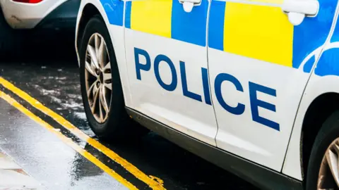 The side of a police car, with "police" emblazoned on it in blue on a white background, parked up on the side of a road which is wet because it has been raining.