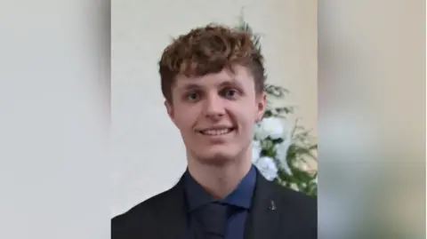 FAMILY HANDOUT Kacper Dudek, a young man with short dark blond hair, wearing a dark suit and blue shirt 