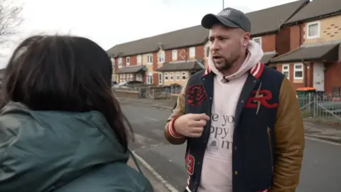 Poet Connor Allen stands in front of some houses. In front of him is an interviewer