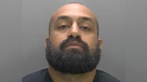 A custody mugshot of Maroof Rahman, who is wearing a black T-shirt and has no expression on his face. He has a shaved head and a bushy black beard.