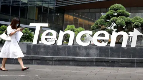 A woman in a white dress, looking at her phone and wearing a face mask, walks outside the headquarters of Tencent in Shenzhen, China.