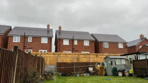 A garden with a row of houses behind it, there is a fence in between the garden and the houses. A washing line can be seen stretching from the right hand side to the left rear corner of the garden. There is a green-painted shed in the right hand corner of the garden.