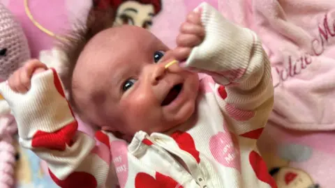 Dolcie-Mae smiling in her cot. She is wearing a top with love hearts on it that say 'I love mum'