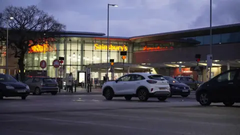 BBC Cars are parked outside the Sainsbury's store on Wilmslow Road in Cheadle. The image is taken at dusk with the inside of the supermarket lit up by artificial lighting. The store's orange logo is prominently displayed in lights.