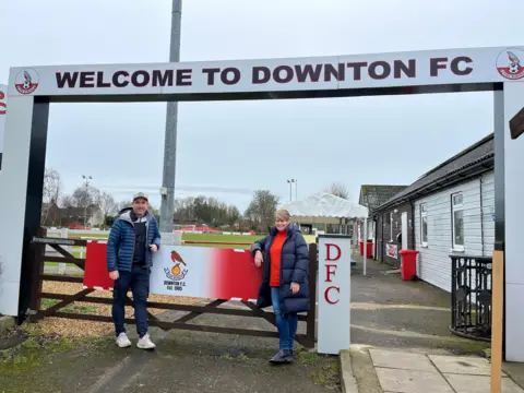 Two people stand in front of the gates to Downton FC. They are smiling at the camera and leaning on the gate. A clubhouse and football pitch are visible behind them. 