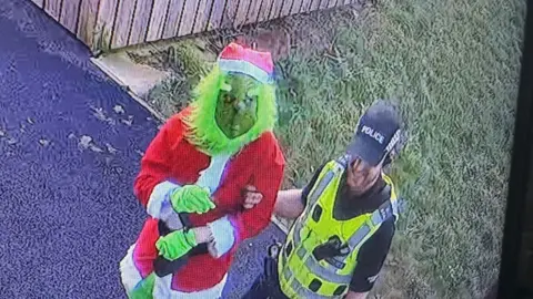 Man dressed in Santa Grinch costume being arrested by police officer in black cap and yellow hi-viz jacket.