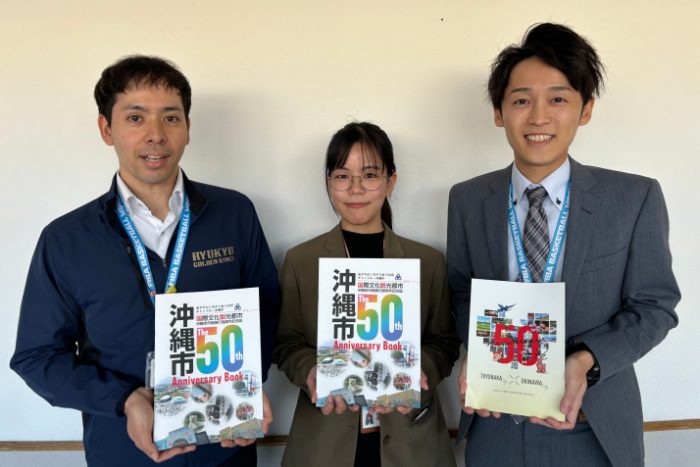 豊中市からの派遣職員で同市の記念誌を手にする沖縄市文化芸能課の久保貴大主事（右）と、沖縄市の記念誌をアピールする秘書広報課の上原由夏さん（中央）、島袋直樹係長＝8日、沖縄市役所