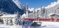 The famous red Bernina Express train passing by the bell tower of Bever alpine village covered with snow, Graubunden canton, Switzerland