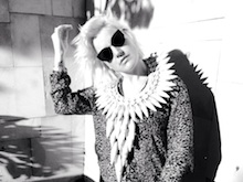 A 30-year-old woman is shown in upper body shot, taken in black and white. She has blonde hair; and wears sunglasses, a leopard pattern shirt and an elaborate shell necklace. Her head is slightly tilted to her right, with her right arm bent at the elbow and her fingers close to her hair. Background walls include some artworks.