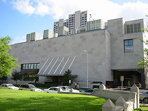 Squat, concrete building, with large drive-through under a wing. A roof for the entrance is attached to the building by angled poles.